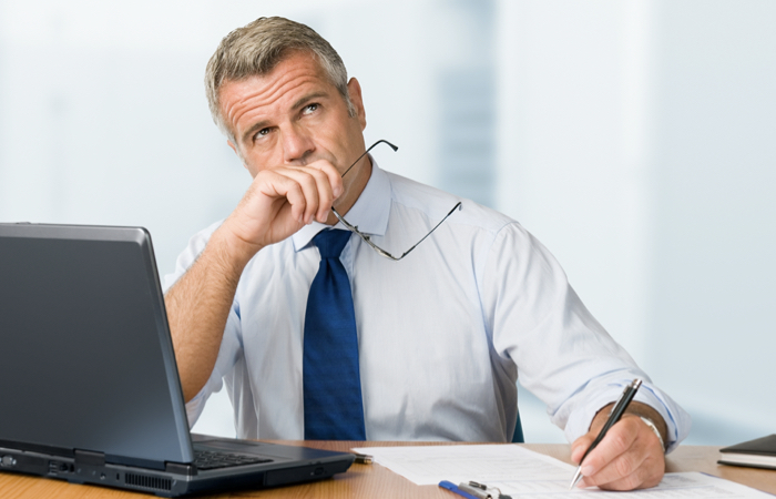 Homem pensativo, vestindo camisa social, gravata e sentado sobre uma mesa com um notebook na sua frente