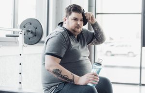 Homem branco com tatuagem na mão direita e visível sobrepeso. Está sentado em um canto de alguma ambiente com uma garrafa de água na mão.