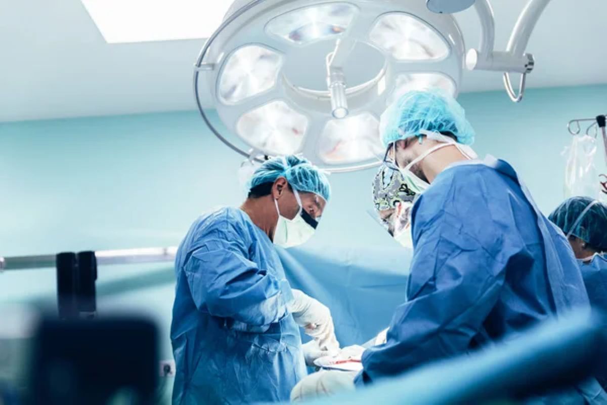 Três médicos em uma sala de cirurgia. Todos estão com roupas azuis e dois com touca da mesma cor. O terceiro usa touca desenhada.