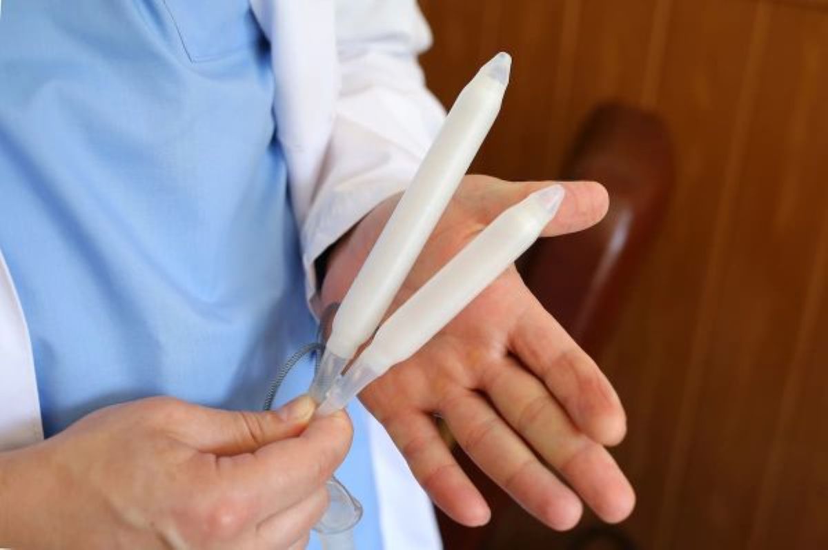 Profissional da saúde com roupa azul e jaleco branco segura uma prótese peniana com as duas mãos.