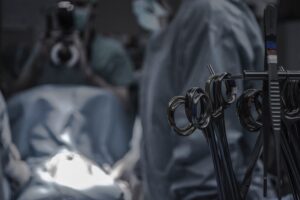 Imagem mostra um centro cirúrgico com amteriais como tesoura em primeiro plano e atrás cirurgiões com roupas cirúrgicas durante a cirurgia de colocação de prótese peniana