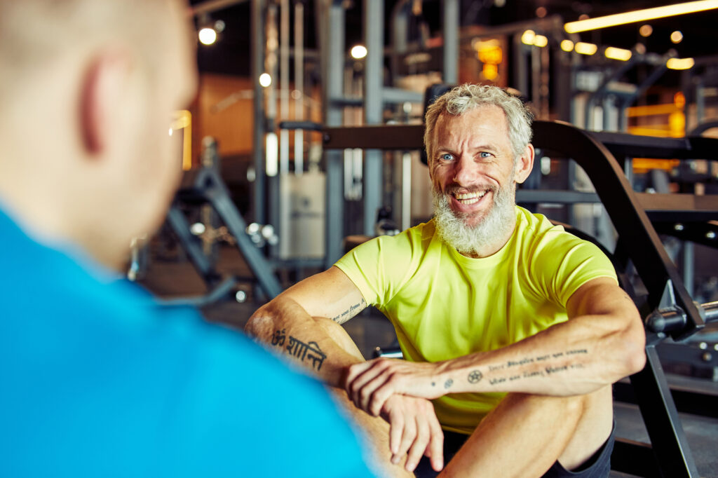 Retrato de um homem feliz de meia-idade discutindo resultados de treinamento com instrutor de fitness ou personal trainer e sorrindo enquanto sentados juntos no chão da academia