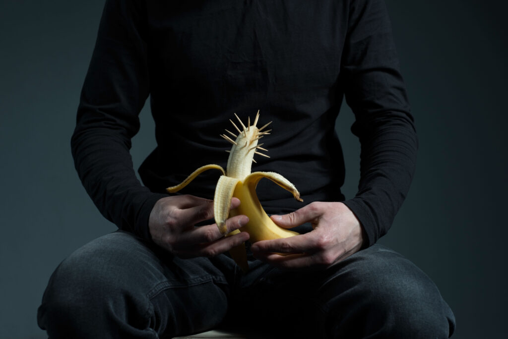 Homem segurando uma banana com vários palitos ao redor da ponta da banana representando dor na glande peniana. 