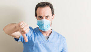 Médico usando máscara com rosto irritado, mostrando o resultado negativo da cirurgia com os polegares para baixo.