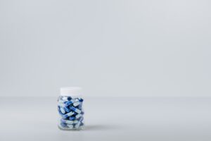 Imagem com fundo branco. Em uma superfície branca há um recipiente com medicamentos de cor azul em seu interior e tampa branca