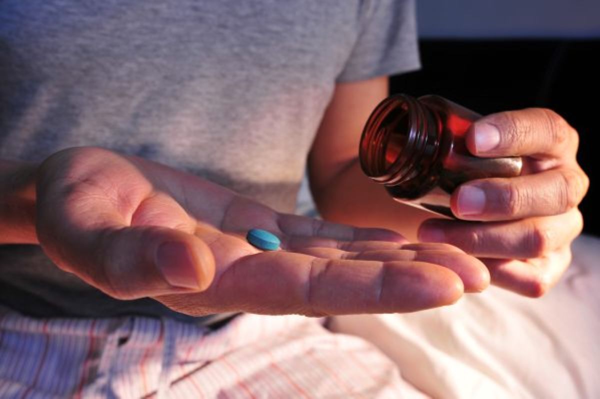 Man in gray T-shirt and light checkered pants sitting on bed holding a brown bottle and a blue pill.