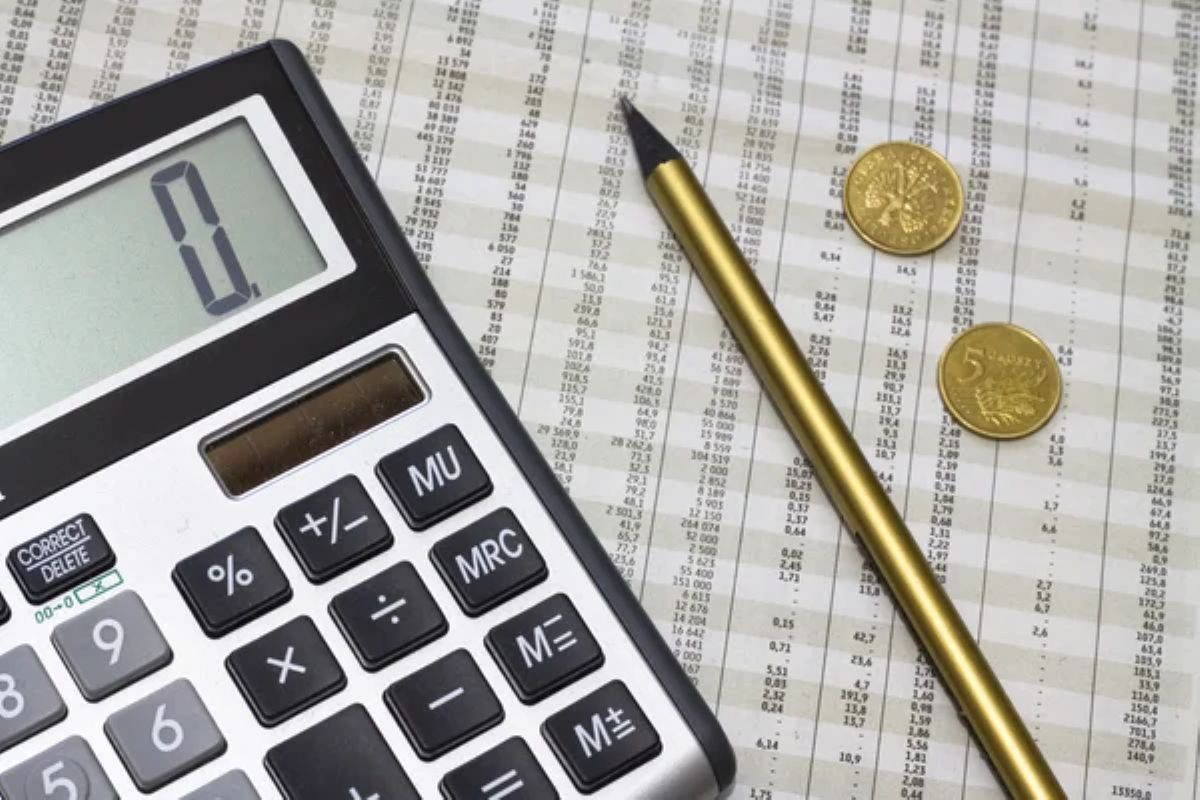 Calculadora sobre uma planilha preenchida ao lado de uma caneta dourada e moedas.