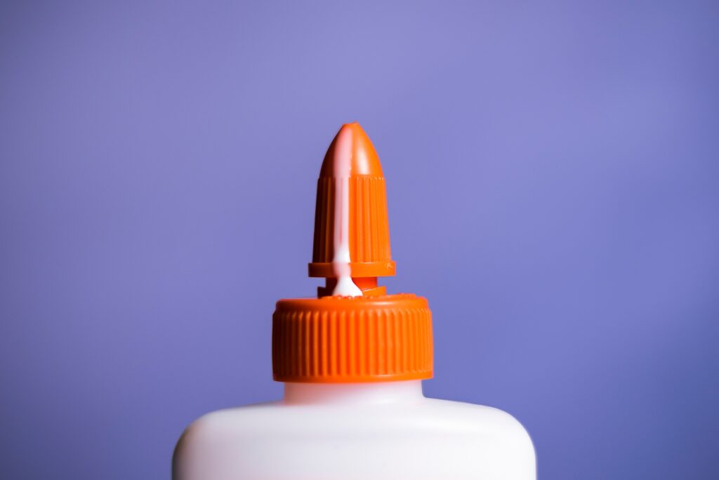 Parte superior de um tubo de cola com líquido branco escorrendo simulando excitação com prótese peniana. A imagem está sobre um fundo roxo.