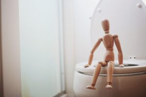 Banheiro com privada branca e boneco de madeira marrom em miniatura sentado sobre ela representando homem com Sling suburetral e esfíncter urinário artificial