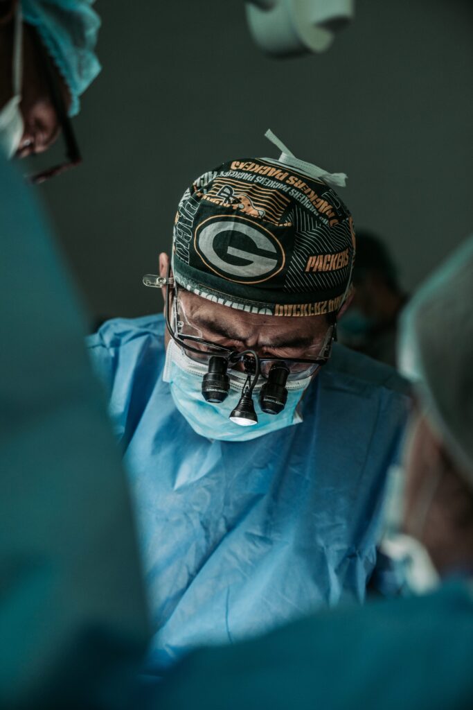 Centro cirúrgico com médico realizando cirurgia de convênio para prótese peniana. Ele usa máscara, óculos especiais, touca e roupas especiais.
