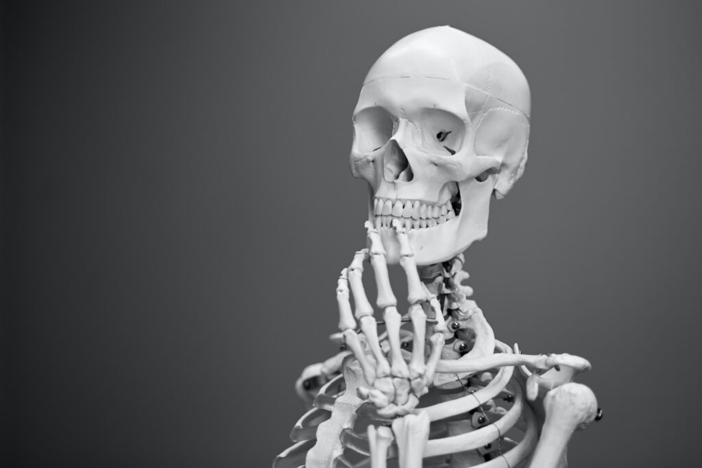 Fundo cinza com imagem gráfica de esqueleto humano mostrando cabeça, ombros e uma parte do tórax, com mão no queixo em pose reflexiva. A imagem mostra o questionamento sobre a calcificação peniana