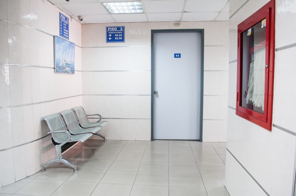 Sala de espera branca, com porta, cadeiras, quadro de avisos e placas indicativas. Ela representa SUS ou convênio para prótese peniana para o tratamento de doenças