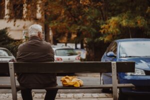 Homem idoso sentado em banco próximo a um estacionamento com carros parados. Ele é branco, tem cabelos brancos e usa casaco e calça em tons de marrom. Ao seu lado há um envelope amarelo. Ele está pensando em envelhecimento e saúde sexual masculina.
