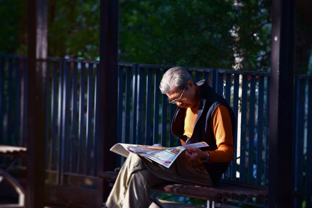 Fundo urbano com grade e árvores e homem idoso sentado em banco lendo sobre terapia para disfunção erétil. Ele veste camisa laranja, calça cáqui e um casaco azul sobre os ombros. Ele está de óculos e tem o cabelo curto e branco.