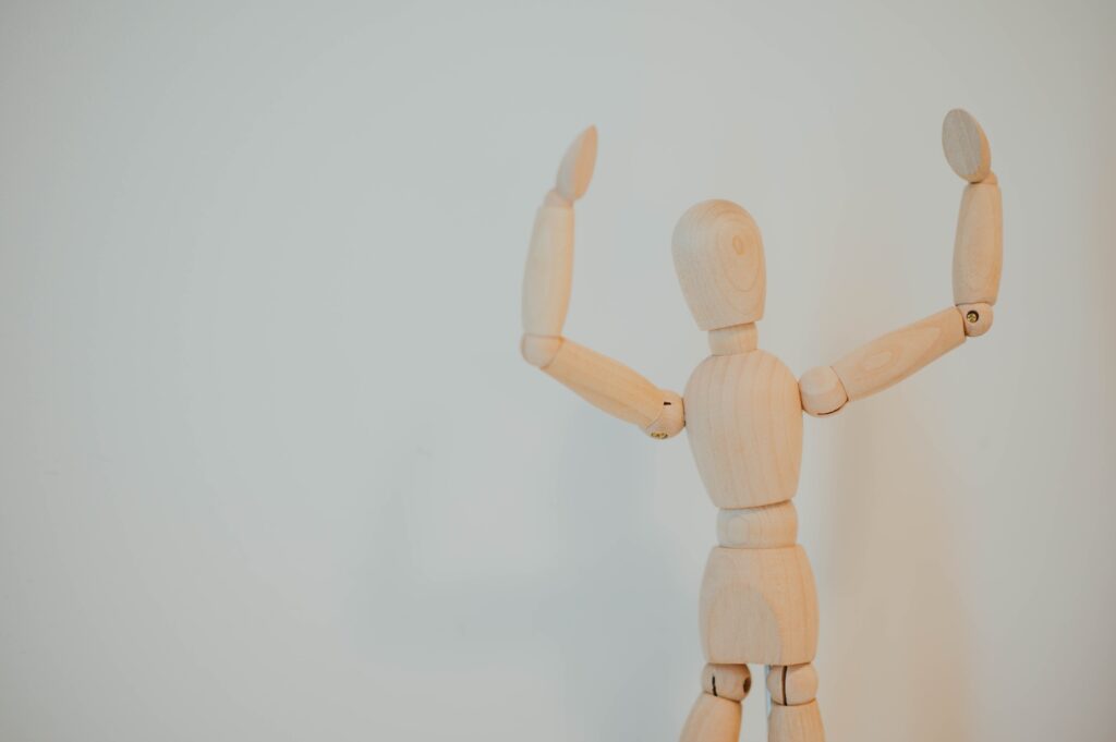 Fundo branco e boneco de madeira clara em primeiro plano com gesto que indica algo grande e simboliza o extensor para prótese peniana