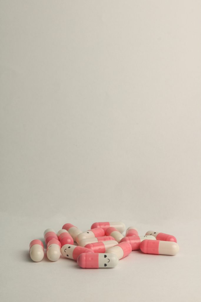 Fundo branco, superfície branca e medicamentos do tipo pílula de cor branco e rosa com rostos delizes desenhados simbolizando efeitos colaterais dos antidepressivos para saúde sexual