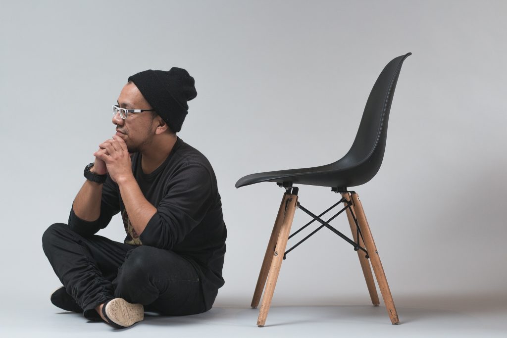 Homem negro com roupa preta sentado no chão ao lado de cadeira preta e marrom pensando sobre a cirurgia de reconstrução peniana