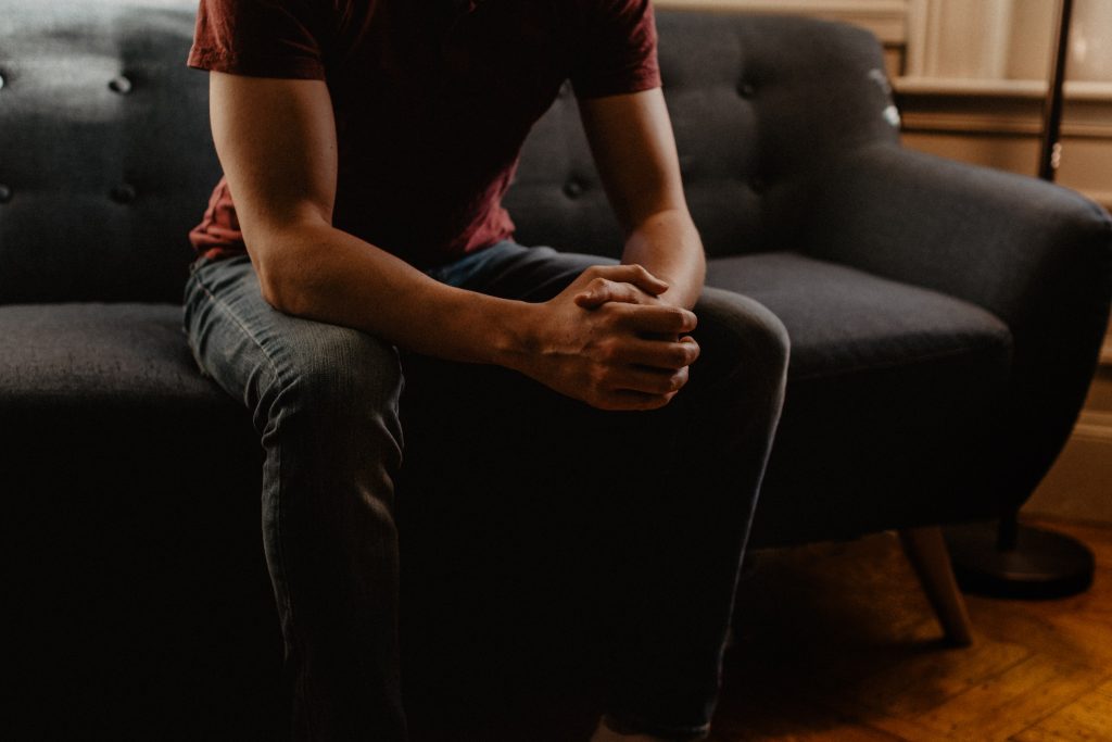 Homem branco de calça jeans e camiseta vermelha sentado em sofá cinza pensando sobre a cirurgia de reconstrução peniana