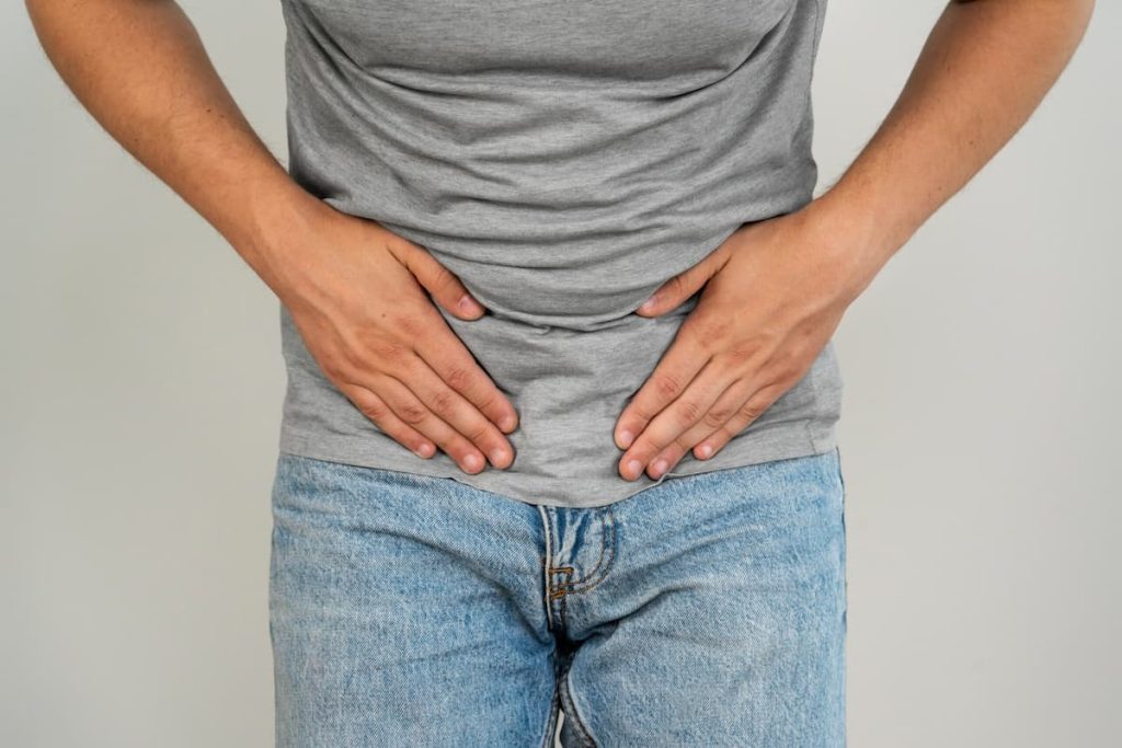 Fundo cinza, homem branco de camiseta cinza e calça jeans da cintura até o meio da coxa. Ele está com as mãos na região genital
