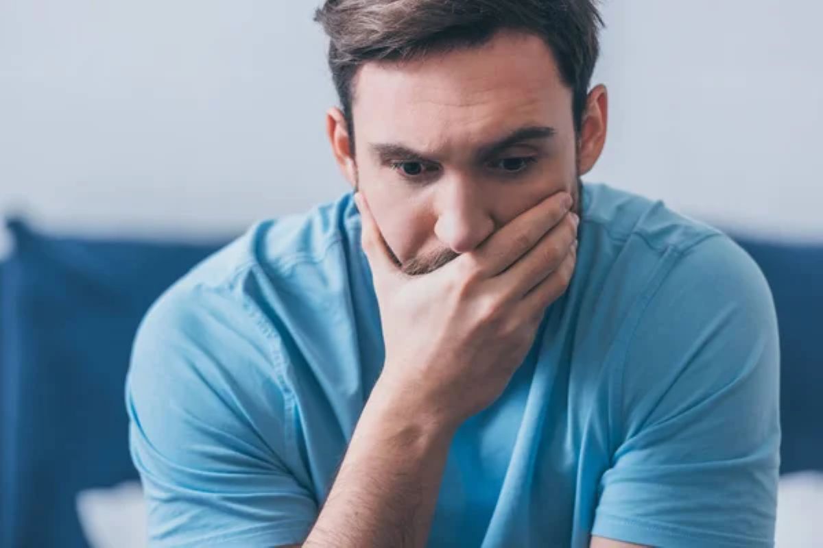 Hombre blanco con cabello y barba castaños, usando una camiseta azul, está sentado con una mano cubriendo su cara. Su expresión muestra preocupación.
