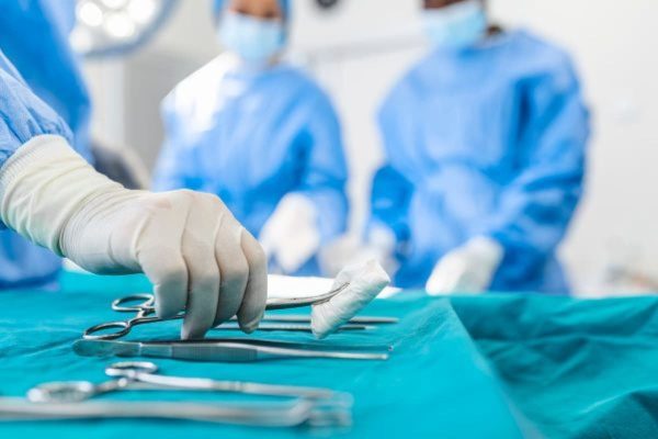 Dois médicos vistos de frente, mas a imagem está borrada. Eles estão de luvas brancas e toucas e aventais azuis. Vemos uma mão com luva branca pegando instrumentos cirúrgicos em uma mesa.