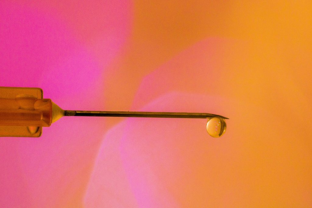 Fundo laranja e rosa e agulha de injeção em primeiro plano com gota transparente simbolizando o que é a harmonização peniana
