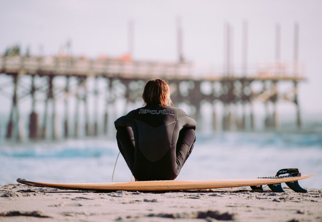 Homem branco com roupa de surf de costas sentado na areia com prancha de surf. Ele olha para o horizonte onde há o mar e um pier. A imagem representa os resultados da harmonização peniana