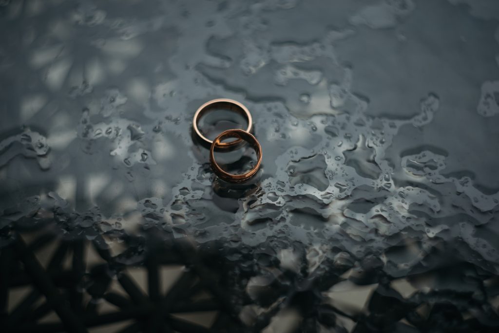 Fundo acinzentado com gotas de água. Sobre ele, duas alianças douradas entrelaçadas simbolizando a parceria de um casal e como ajudar o marido com Peyronie
