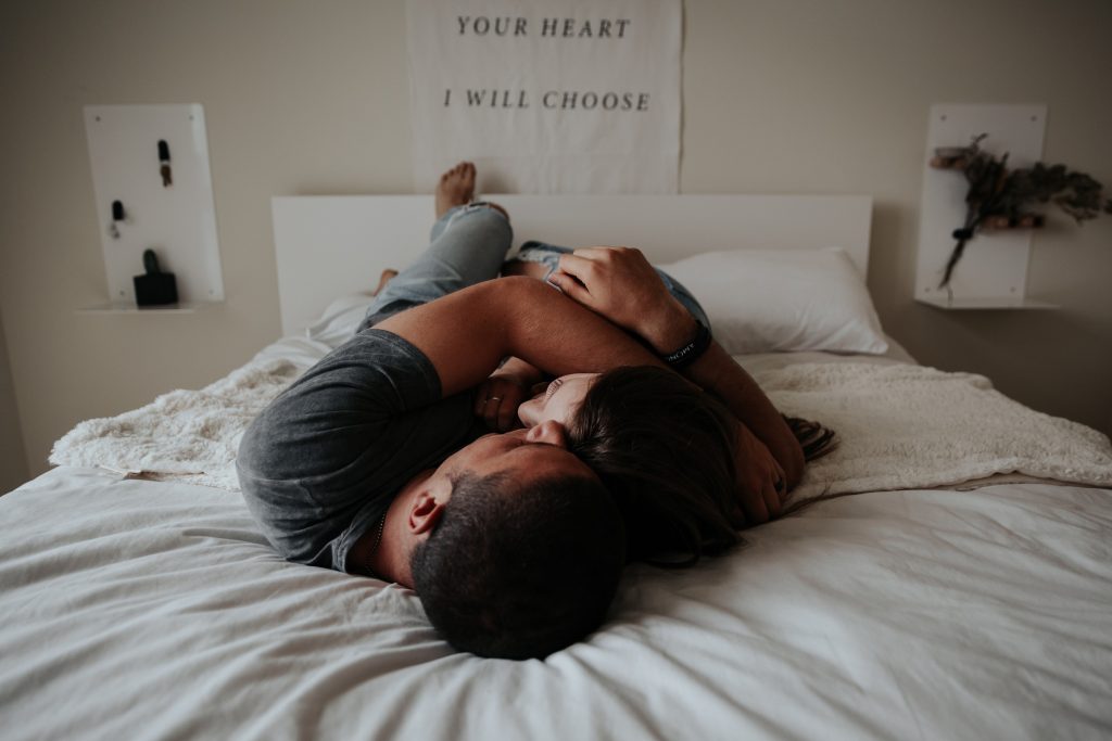 Quarto com paredes, quadros e cama brancos. Em cima da cama há um casal branco abraçado e se acariciando simbolizando a importância da sexologia.