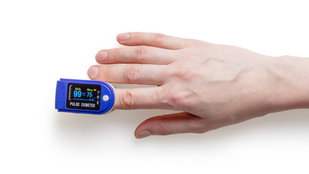 Fundo branco, mão de pessoa branca e aparelho de medir pressão instalado no dedo para aferir a pressão, um dos problemas de saúde que o medicamento atenolol trata