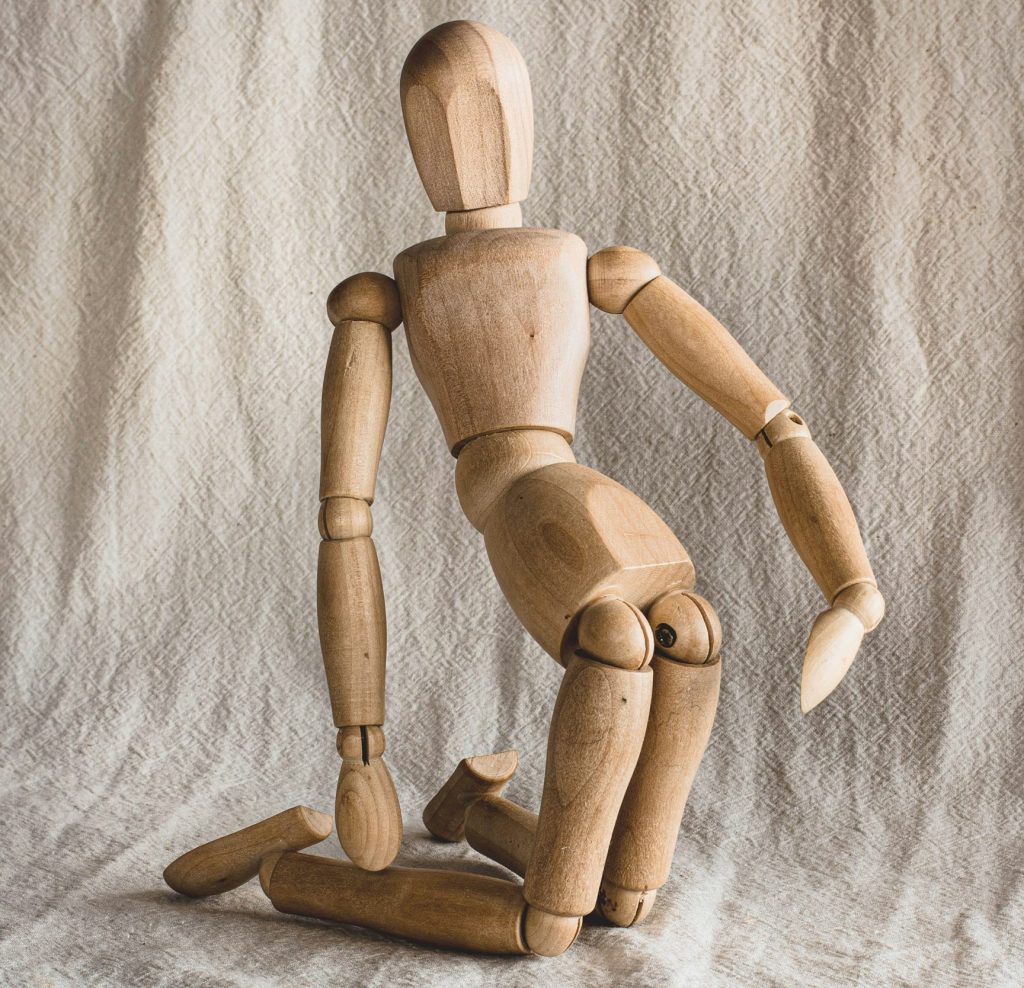 Boneco de madeira clara de joelhos em superfície de tecido de algodão cru projetando o quadril para frente simulando as melhores posições sexuais para quem tem prótese peniana