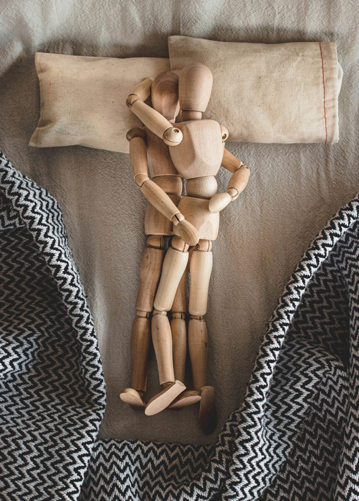 Dois bonecos de madeira clara abraçados deitados sobre superfície cinza com travesseiros brancos pequenos e tecidos com estampa azul nas laterais representando as posições sexuais para quem tem prótese peniana