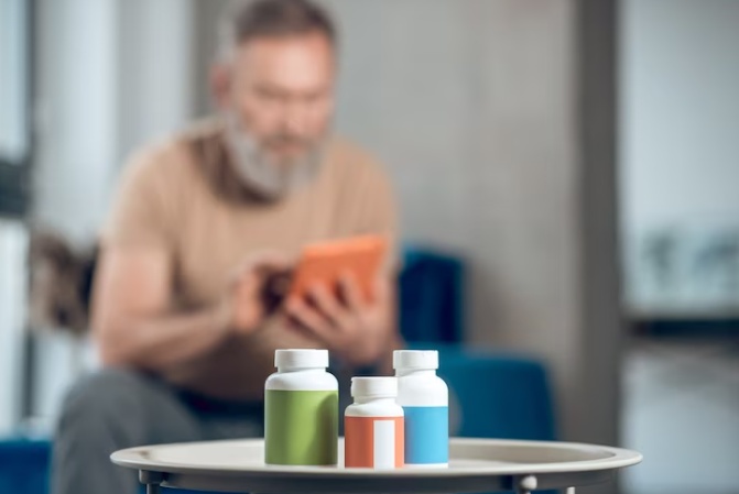 Olanzapina efeitos colaterais: um homem sentado surge no fundo da imagem, desfocado. Na parte central, há três vidros de medicamento sobre uma mesa.