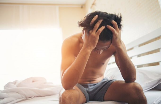 Homem sentado na beira da cama com as mãos na cabeça, o que dá a sensação de que ele está preocupado., referenciando um homem que sofre de disfunção erétil e hipertensão. Ele está de short azul e sem camisa.