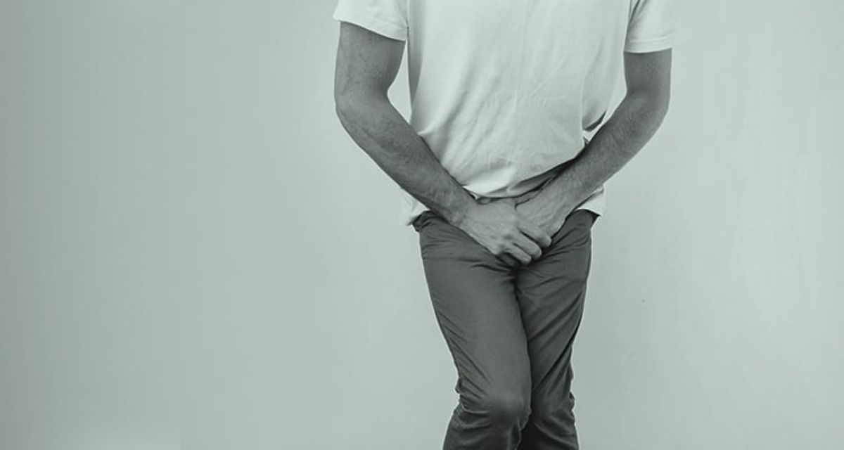 Black-and-white photo of a man wearing jeans and a white shirt with both hands over his penis.