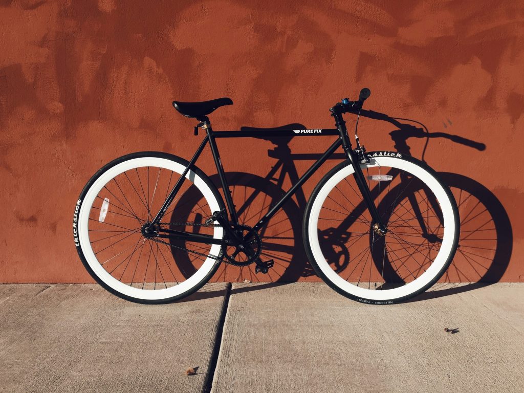 Calçada cinza, muro marrom alaranjado e bicicleta preta e branca encostada com o sol fazendo sombra no muro representando prótese peniana e bike
