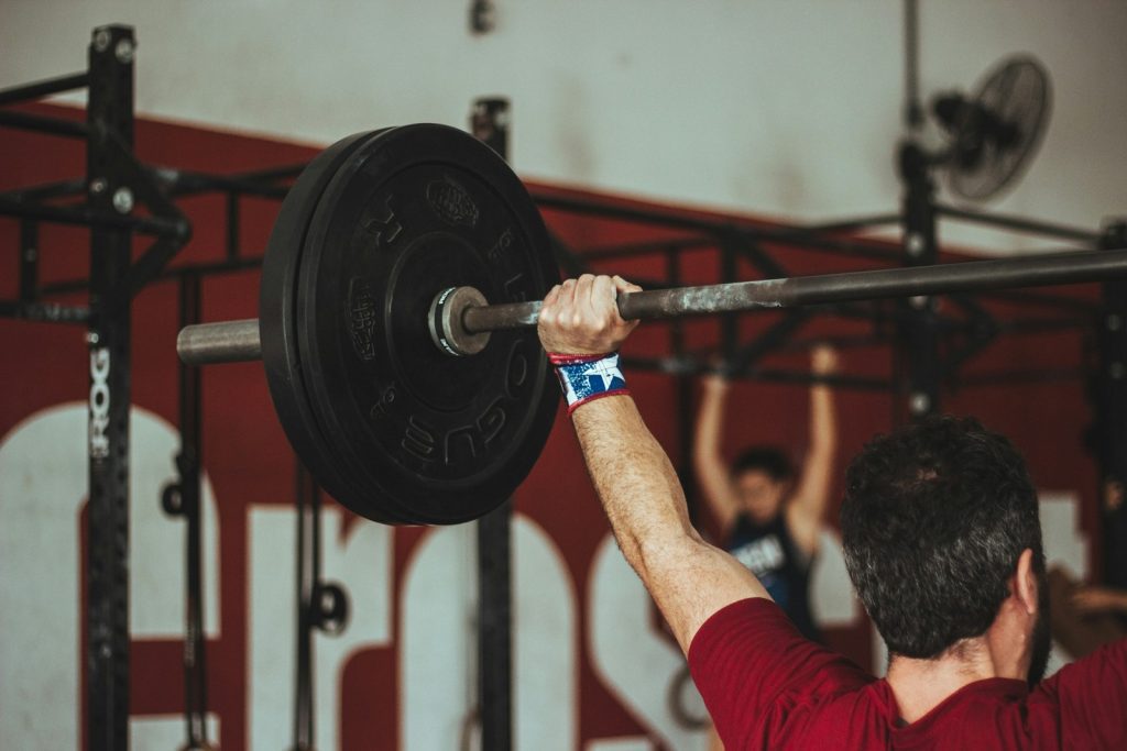 Fundo de academia do tipo crossfit e em primeiro plano homem branco com camiseta vermelha, com vista apenas para o braço esquerdo com pulseira azul, erquendo uma barra com um halter preto pesado representando reposição hormonal masculina e a testosterona