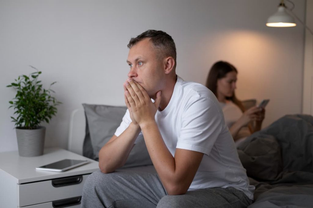 photo of a man sitting on the edge of the bed with his hands over his mouth, looking forward thoughtfully, and a woman sitting in the background using a cellphone