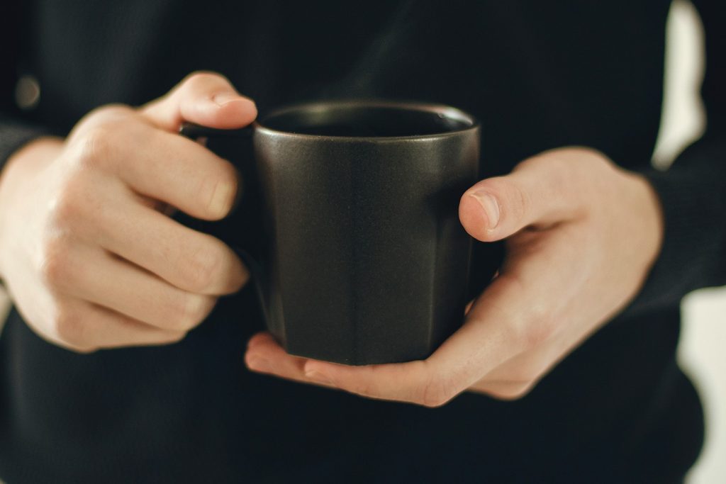 Visão do tronco de homem de blusa preta com mãos à frente do corpo segurando caneca com café, um dos alimentos bons para próstata