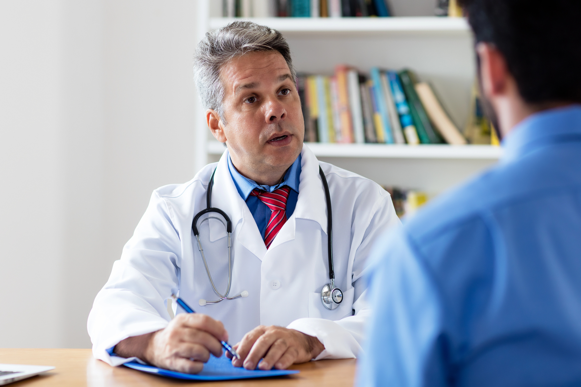 médico idoso de jaleco branco, camisa azul e gravata vermelha conversando com um paciente de costas de camisa azul e moreno, sentados em um consultório com estante de livros e mesa, com prontuário para anotações durante a consulta de prostatite