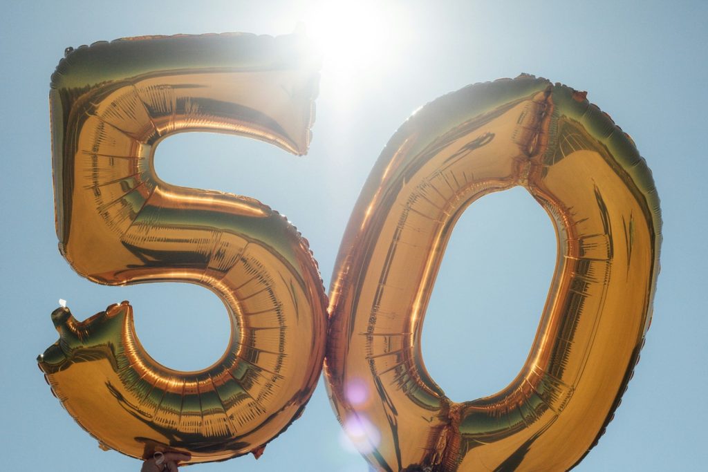 Céu azul, sol na parte superior com muita luz, estourando a imagem, e dois balões dourados inflados com os números 5 e 0, formando 50, que simbolizando o sexo depois dos 50 anos