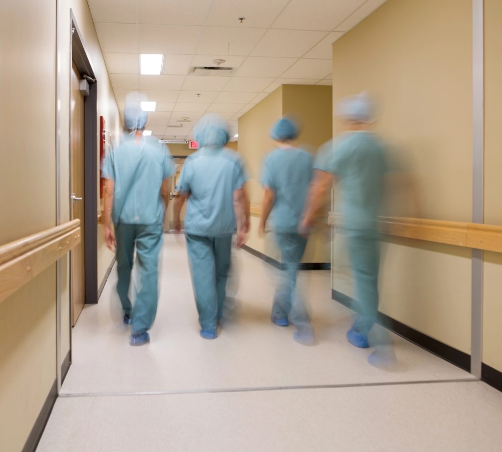 Equipe médica com quatro pessoas de costas e roupas azuis com touca caminhando no corredor bege do hospital indo aplicar os princípios geométricos na curvatura peniana