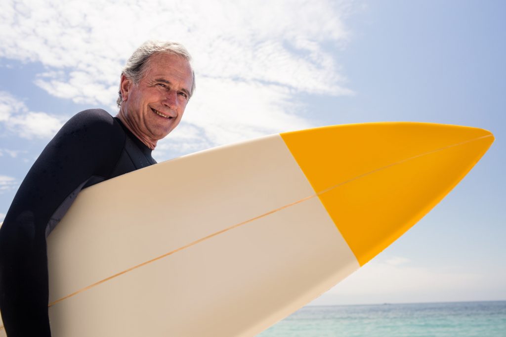 Cielo azul con nubes y mar de fondo; hombre mayor con traje de neopreno negro sostiene una tabla de surf blanca y amarilla, ilustrando beneficios del sexo después de los 60