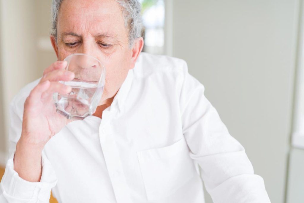 Fondo borroso con pared blanca y ventana, y frente a él, un hombre blanco con camisa blanca bebiendo un vaso de agua, representando si un hombre de 70 años puede tomar Sildenafilo