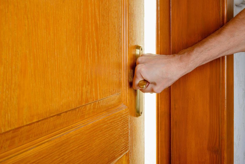 Brown wooden door being opened by a white man's hand on a golden doorknob, revealing a white background on the other side