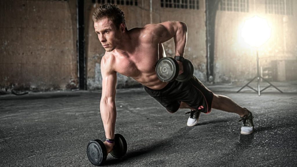 man in a warehouse holding a dumbbell in one hand and another dumbbell that is off the ground in the other hand