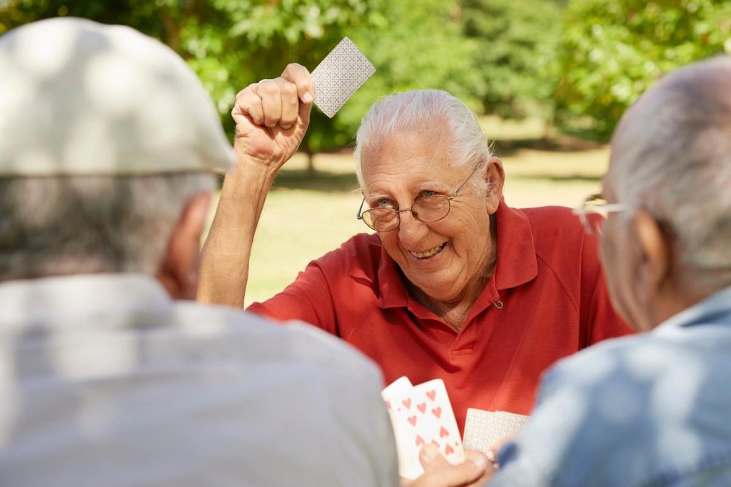 Três idosos, brancos, de cabelos grisalhos, sendo dois de costas parcialmente visíveis e um de frente , com óculos, com uma carta de baralho na mão e expressão contente em uma praça representando a saúde do homem ao longo da vida