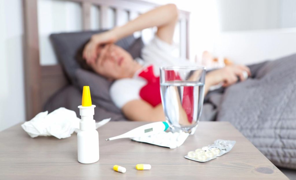 White man in a white t-shirt lying on a wooden bed on gray sheets appearing to have compromised men's health, hot water bottle in his lap. In front, there is a bedside table with a glass of water and medicines.