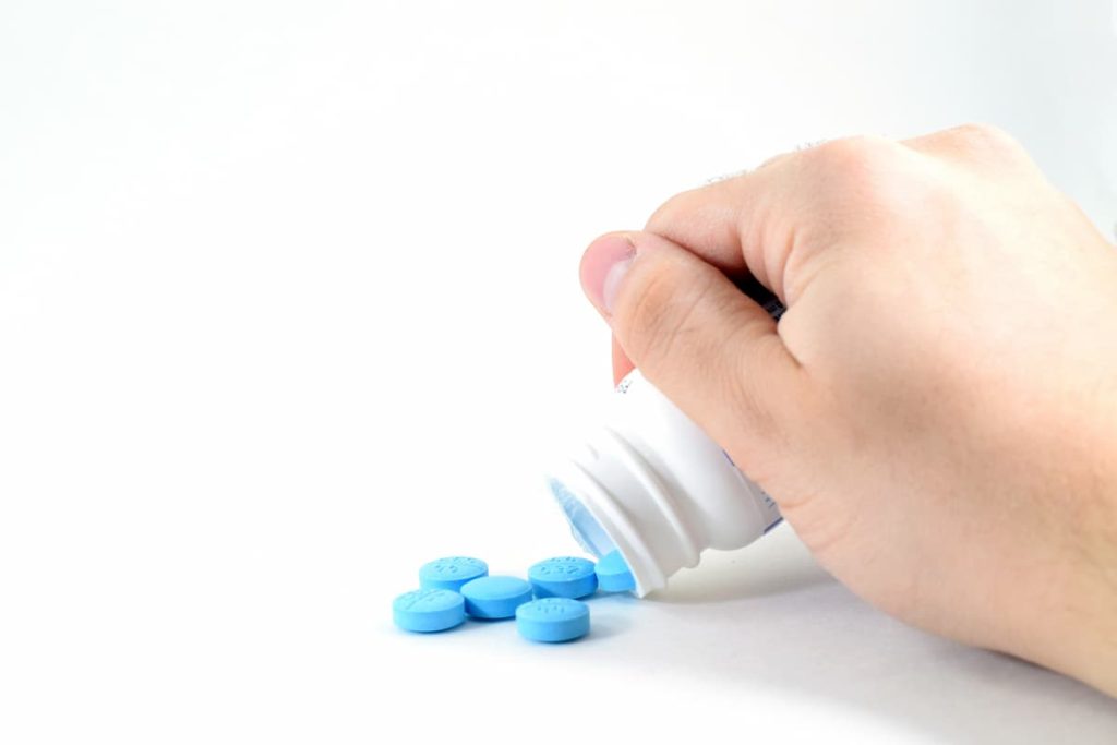 superfície branca sob remédios azuis representando Para que serve citrato de sildenafila, com mão de homem branco despejando os comprimidos de um pote branco