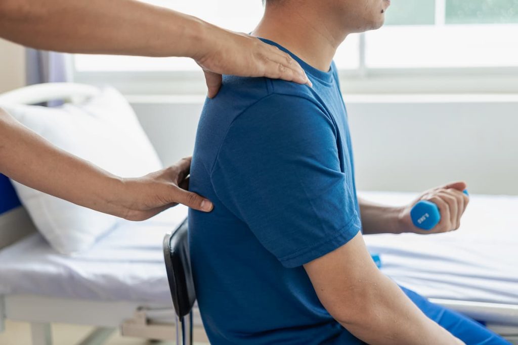 Hombre blanco con camiseta azul sentado en una silla haciendo ejercicio de fisioterapia en la salud del hombre con mancuerna azul y profesional de salud blanco con la mano en la espalda del paciente